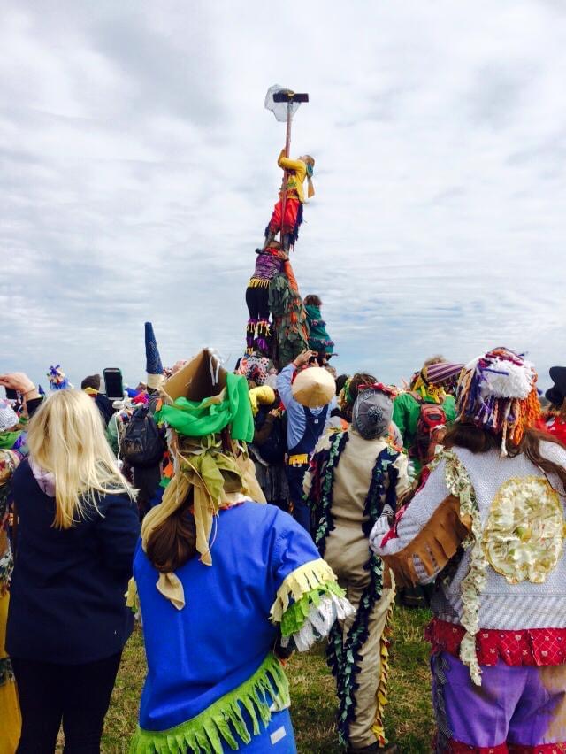 a crowd surrounds a man climbing a tall greased pole
