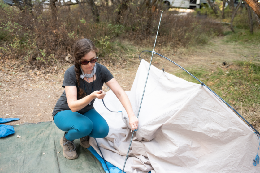 This is a photograph of a Caucasian woman in her 30s setting up a small dome tent while camping outdoors on a summer day in Black Canyon of the Gunnison national park in Colorado, USA.