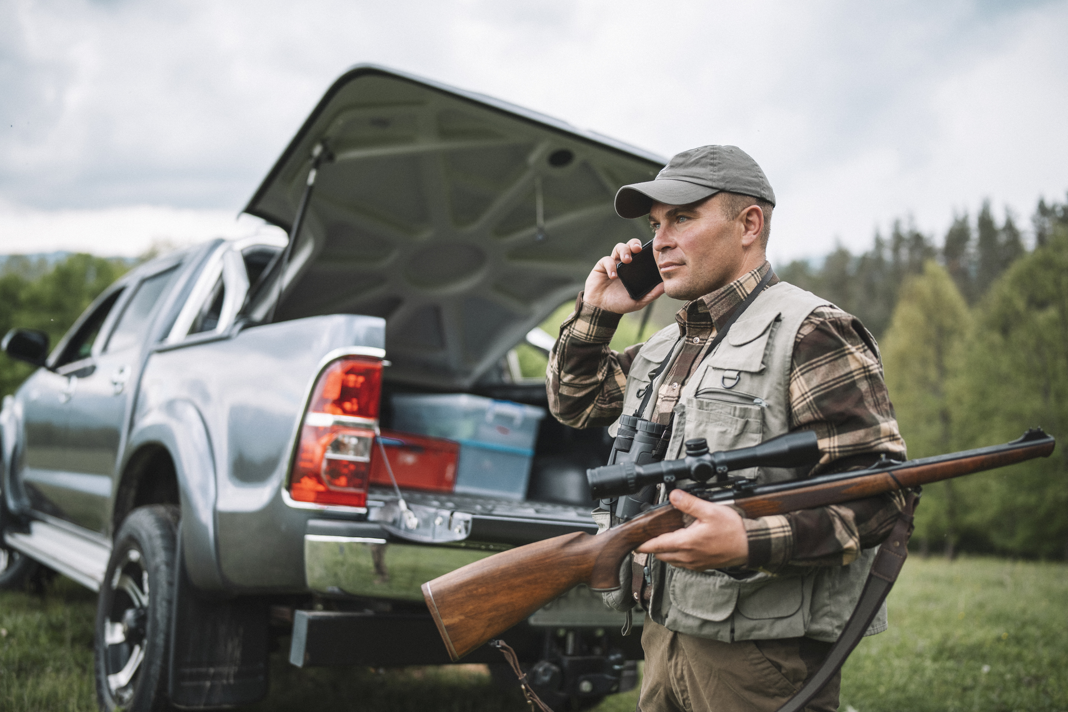 A hunter on a phone and holding his gun in front of his pickup truck.
