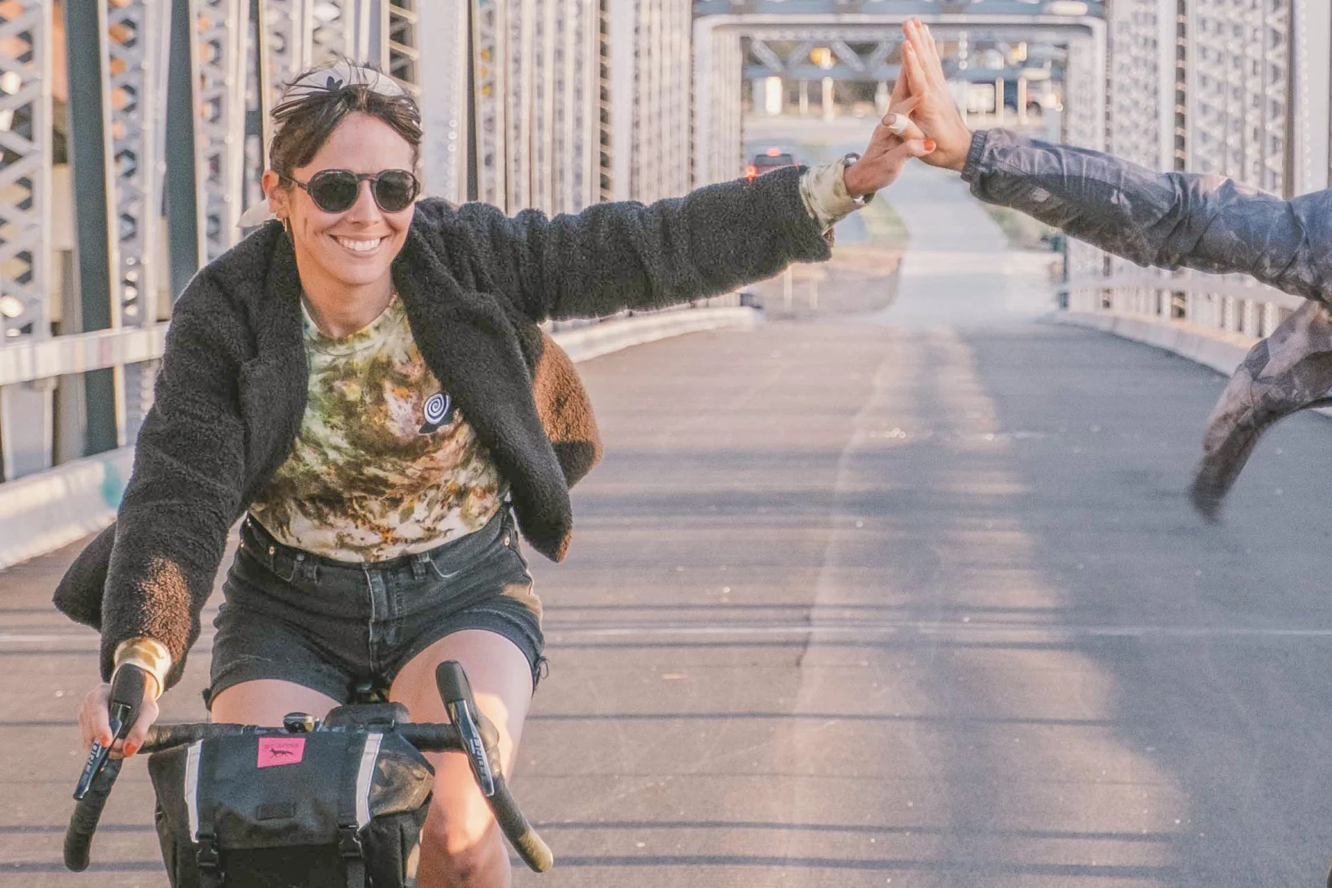 Someone wearing Ombraz sunglasses while biking and high-fiving a nearby cycling friend.