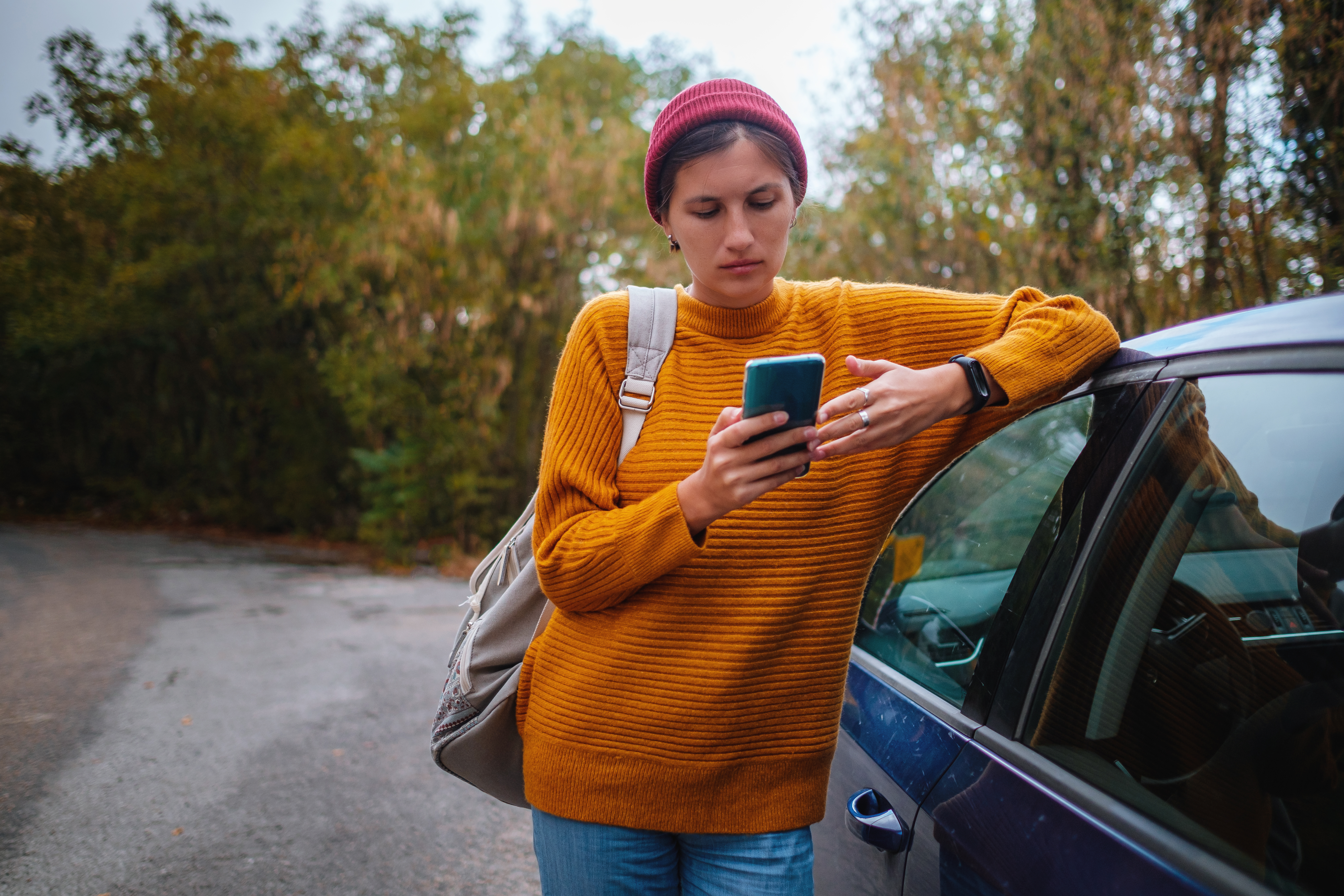 Woman beside a car and looking at smart phone surrounded autumn forest in mountains. Portrait of romantic hipster female, Warm autumn weather, calm scene. Wanderlust photo series.