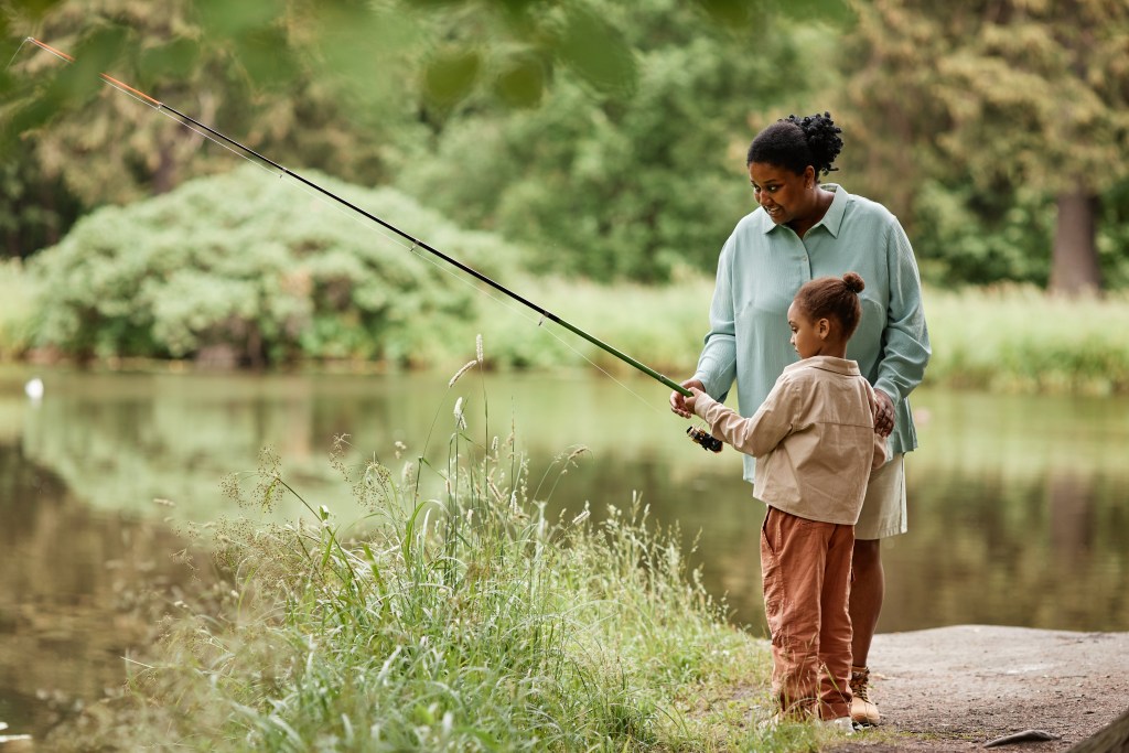 Side view portrait of caring black mother teaching little girl fishing by beautiful forest lake, copy space