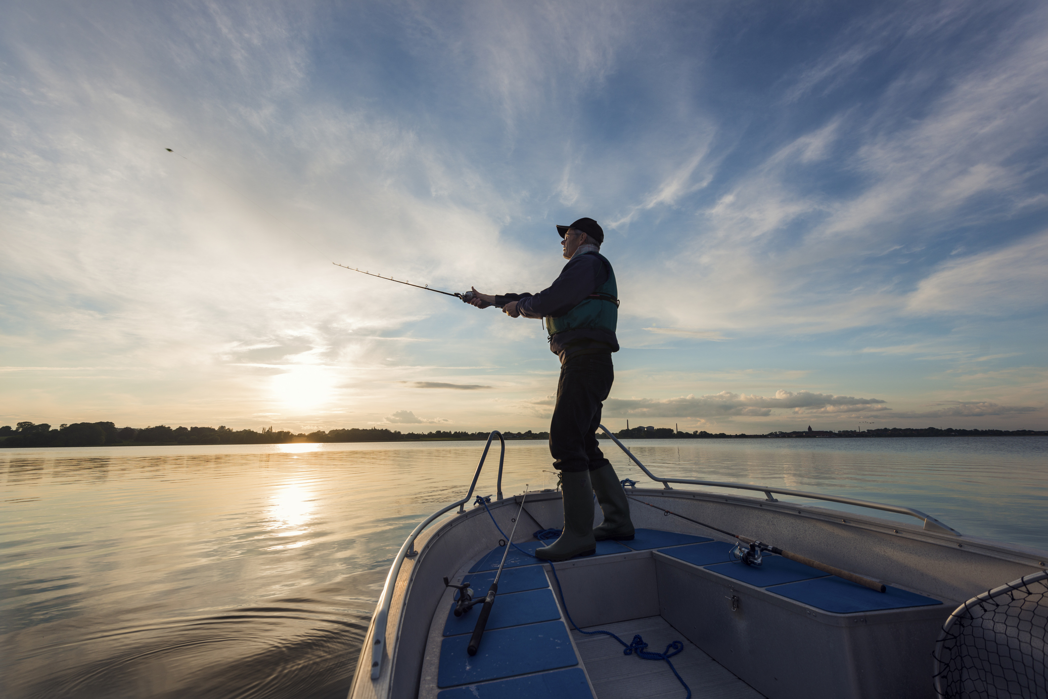 Portrait of a 65 year old fisherman casting out his fishing line into the water from his boat into a flat calm sea. Horizontal format photographed against a setting sun with lots of copy space in the sky. Photographed on Stege Nor on the island of M&oslash;n or Moen in Denmark.