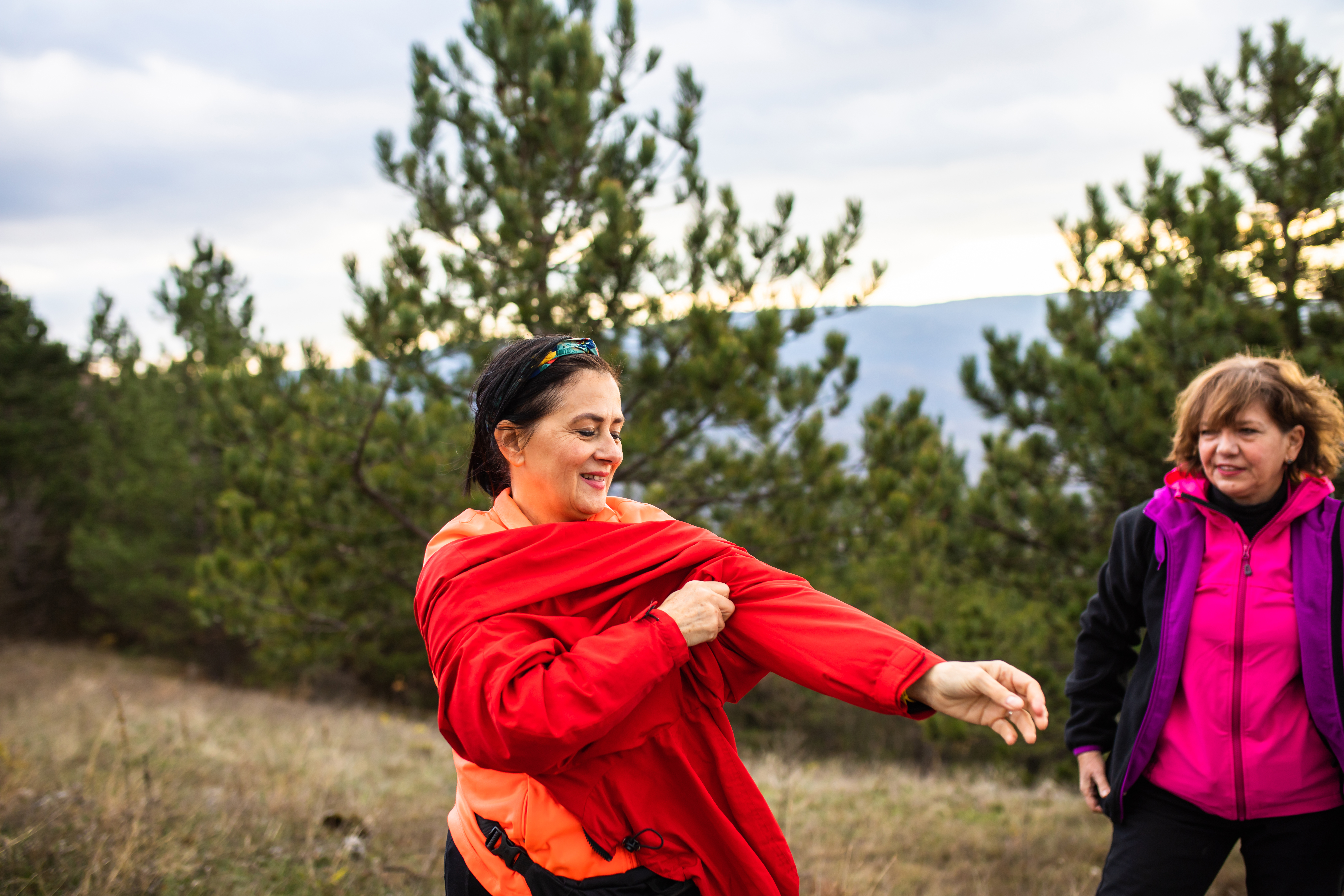 A mature woman is putting on more layers of clothes while hiking on a cold autumn day