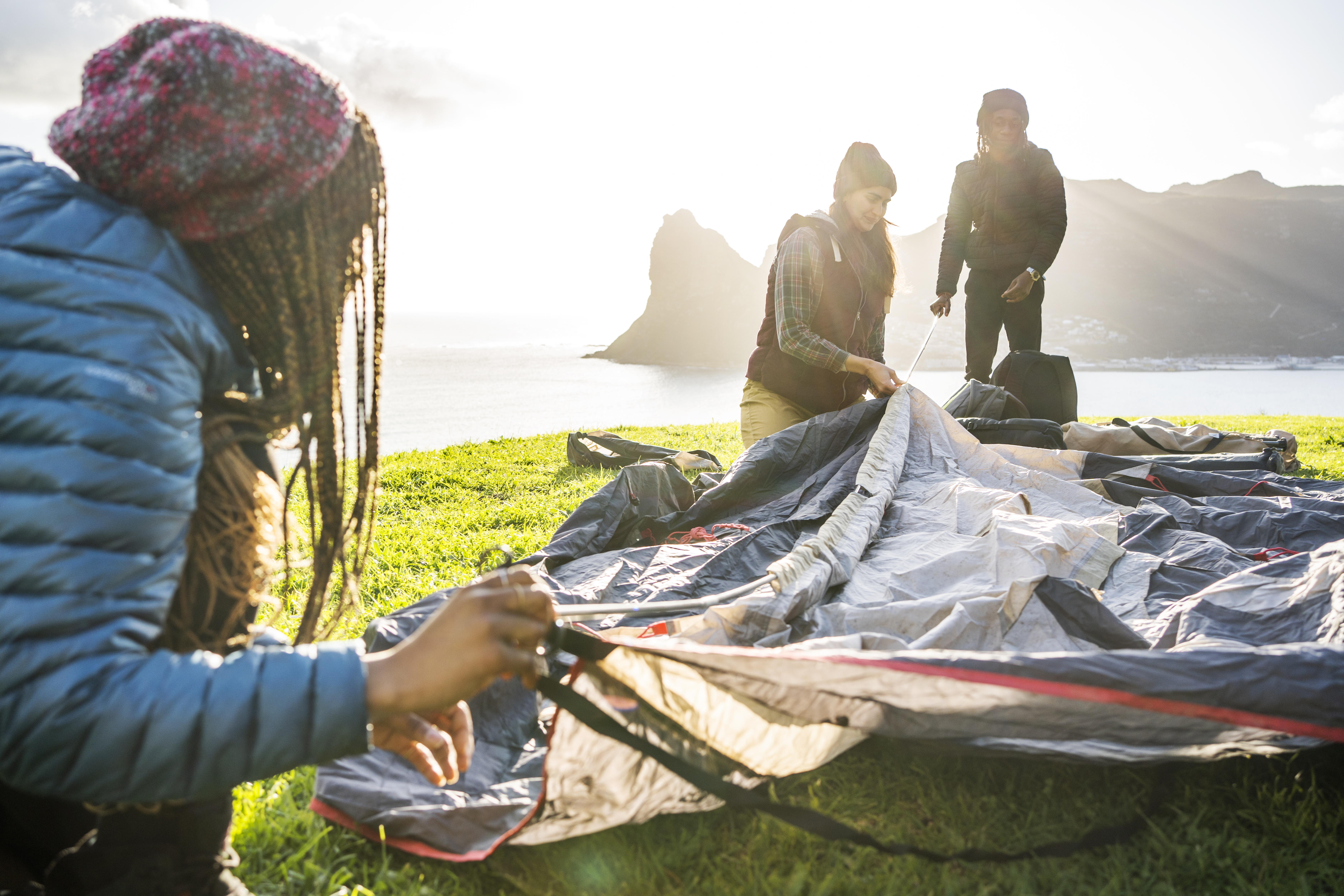 Diverse group of young friends lining up poles while setting up their tent overlooking the ocean during a camping trip together 