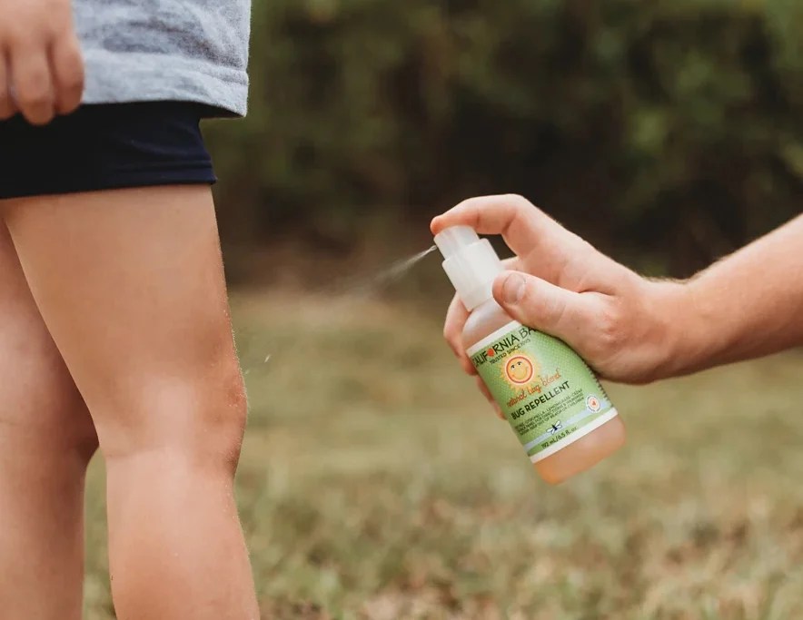 Adult spraying California Baby Natural Bug Blend Repellent onto child's leg