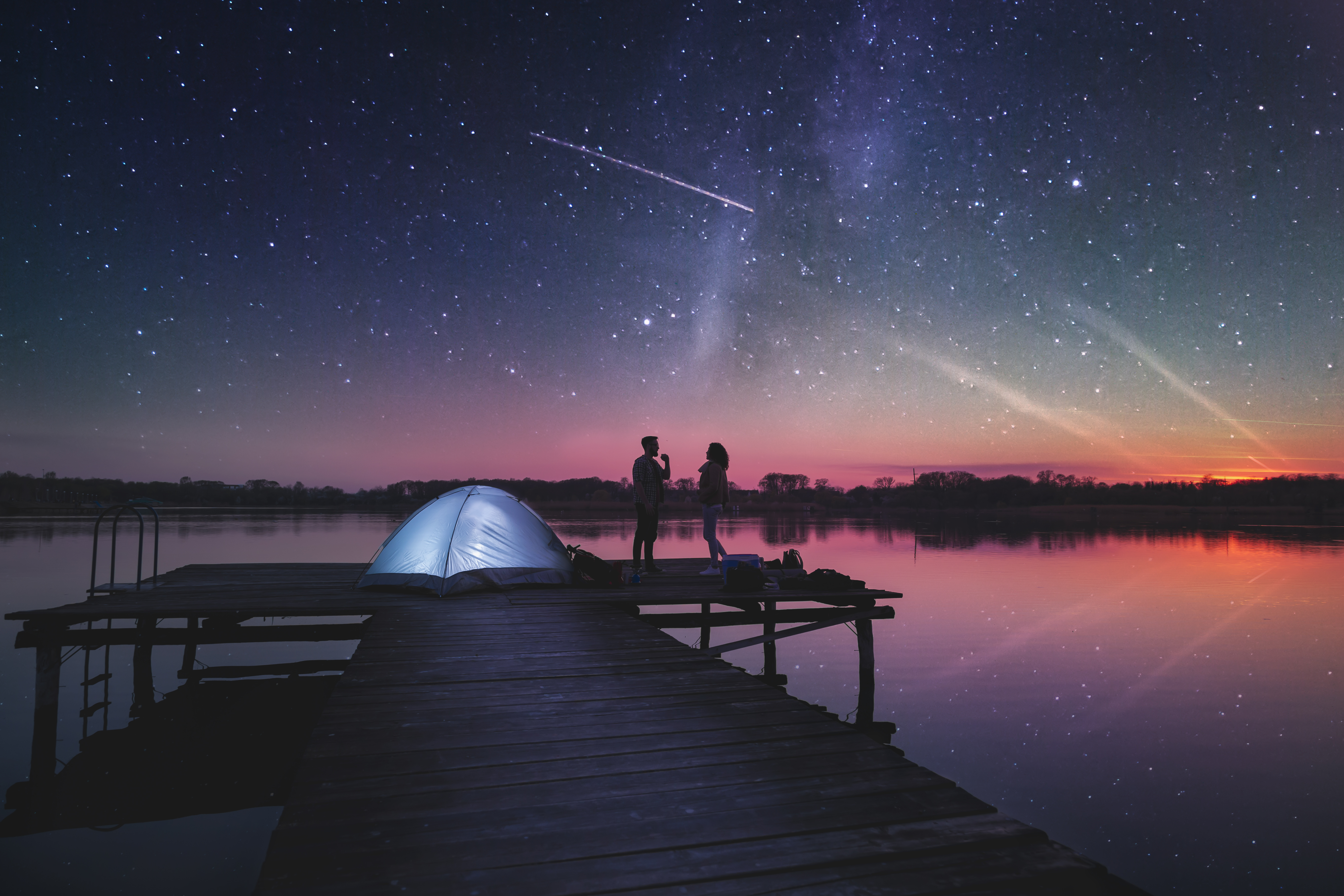 Couple camping on lake docks at dusk, standing next to a tent enjoying beautiful starry night sky
