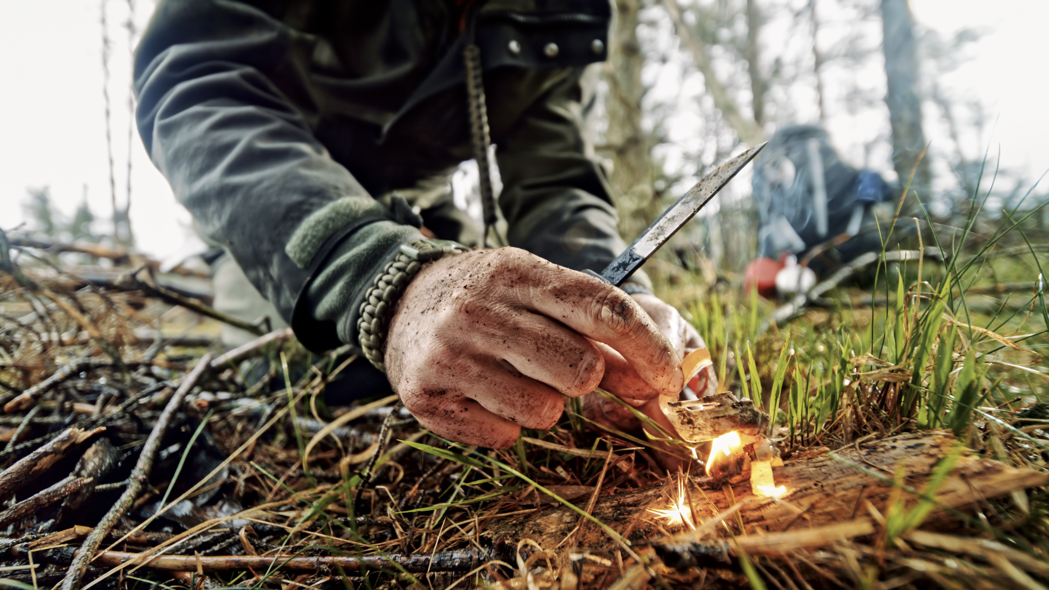 Close-up of man's hands igniting fire using ferrocerium rod and knife in forest.