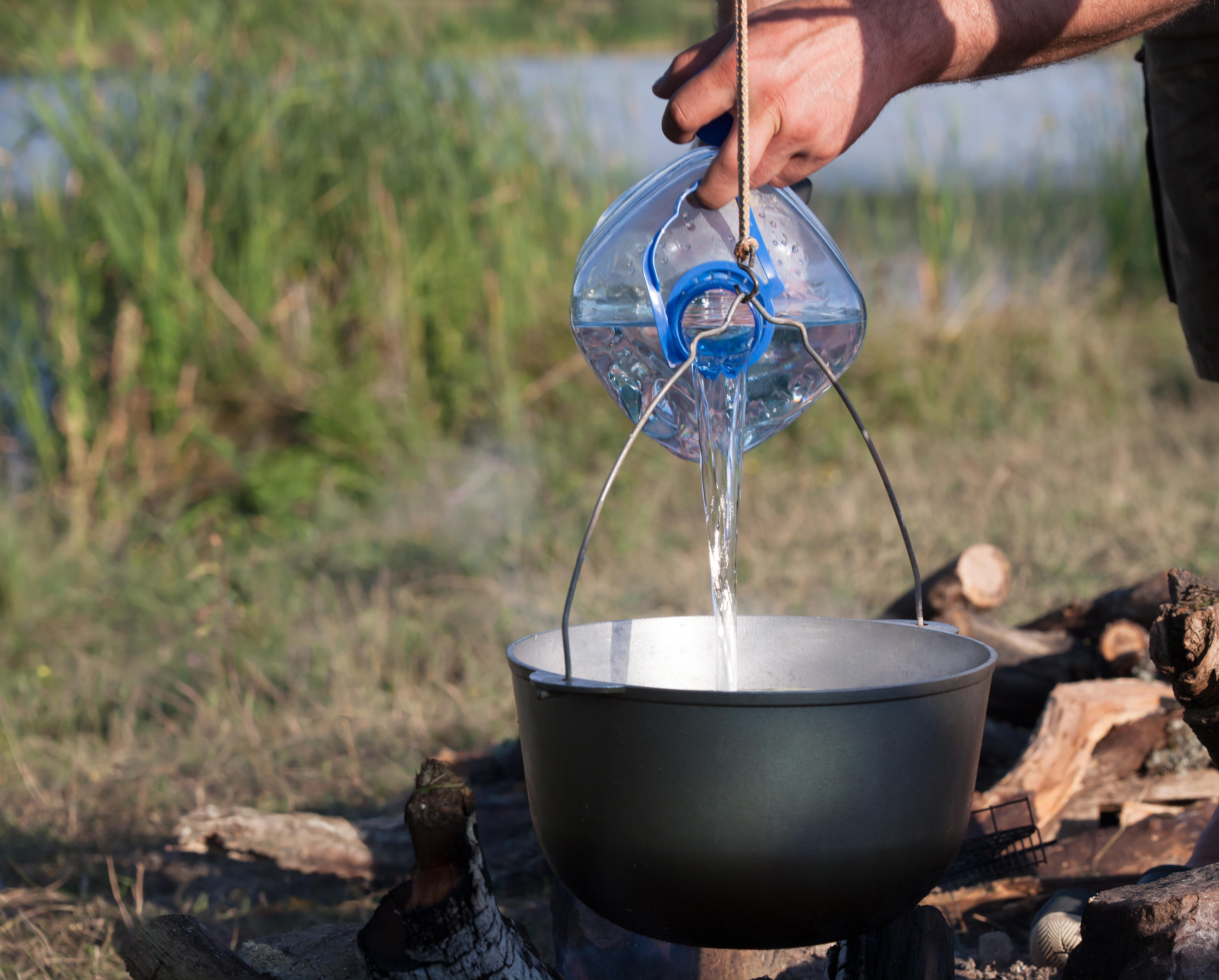 Adding water from a big plastic bottle in black cauldron on open fire, camping meal