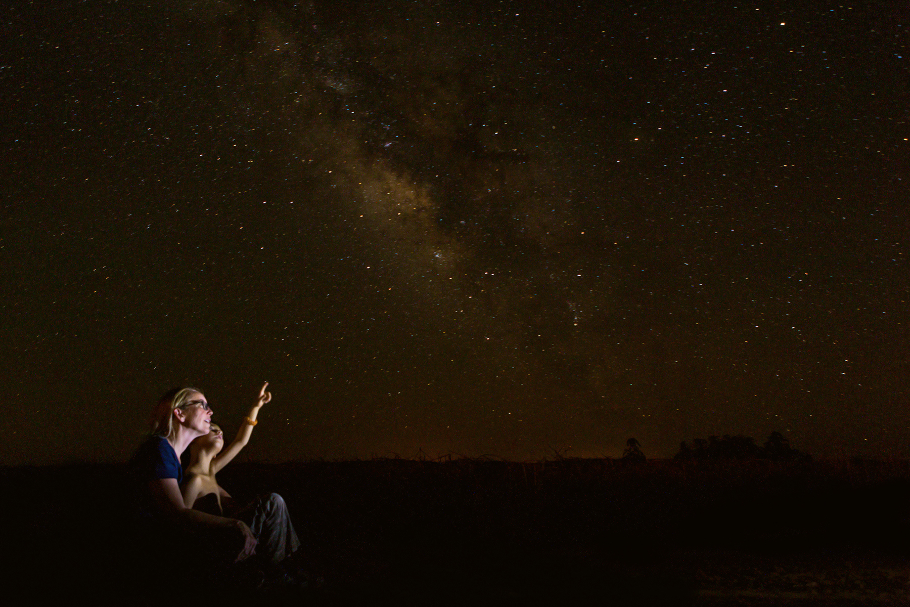 Mid adult Caucasian mother is sitting outdoors with her young son. Boy is sitting in mother's lap while they look up into the night sky. They are star gazing and studying constellations. Little boy is pointing to moon or stars in clear night sky.