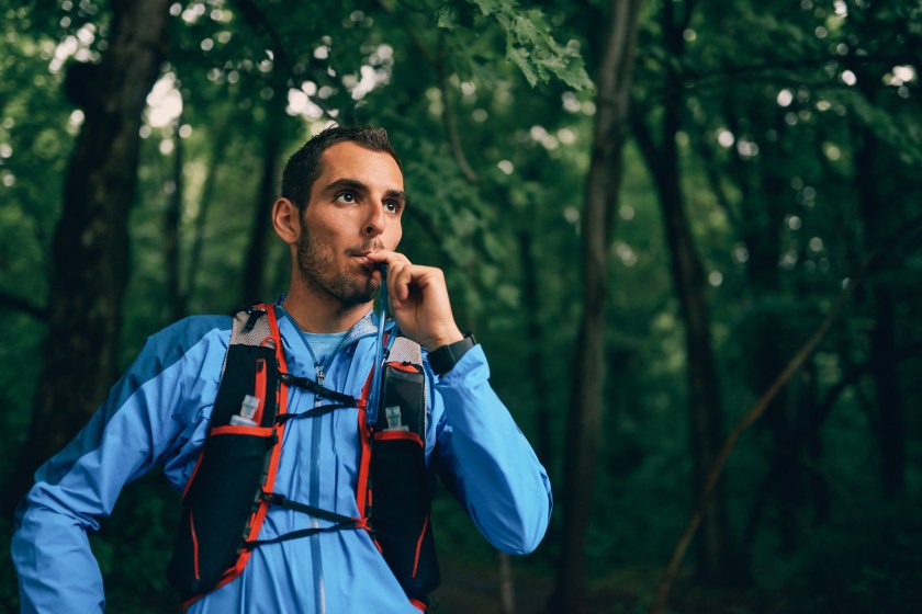 Portait of a competitive, athletic millennial man drinking water from a hydration pack before running off road outdoors through the woods on a trail in the afternoon wearing sportswear.