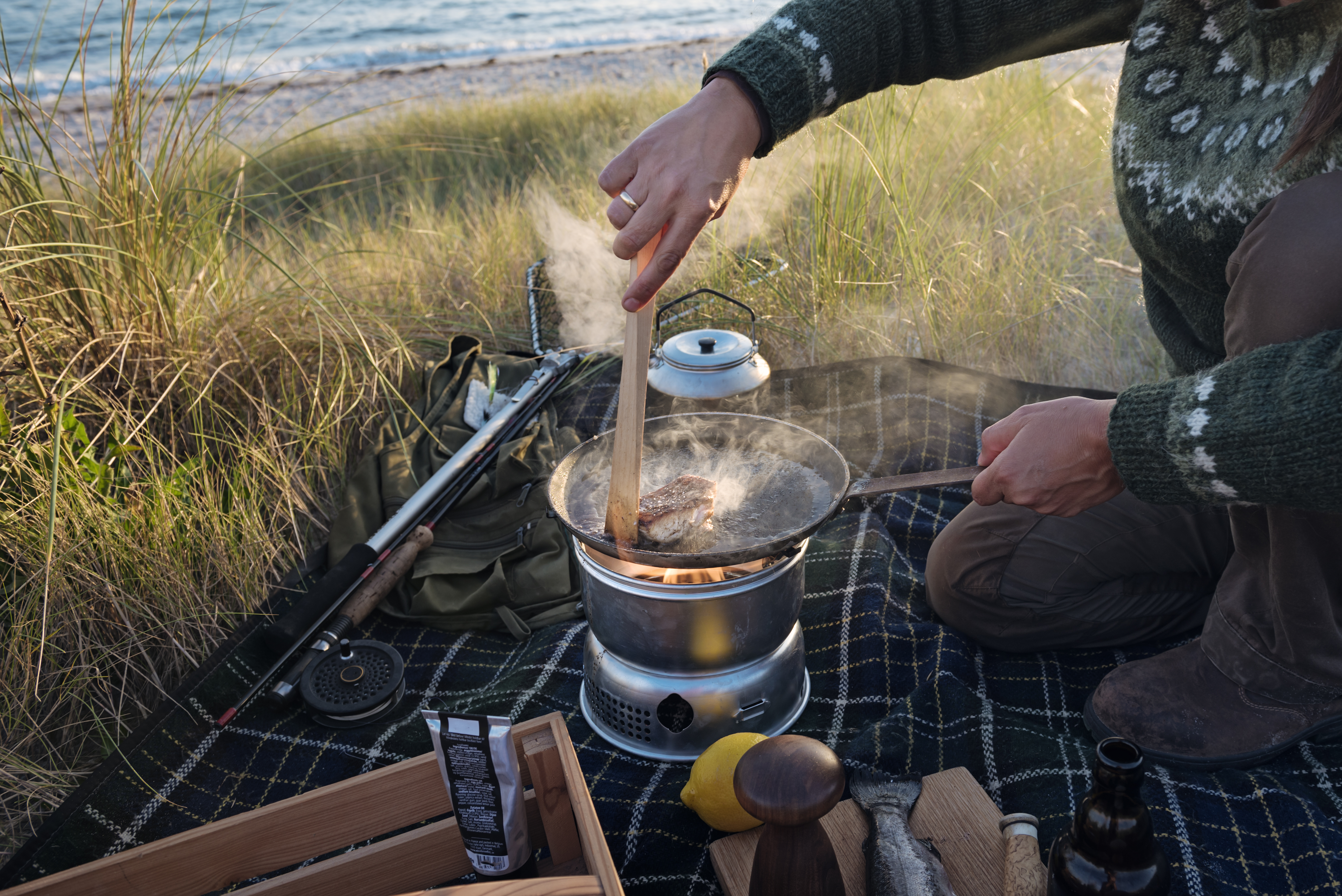 Fisherman cooking her catch at the beach. Female fisherman preparing a filled piece of fish she caught earlier. She is using a gas camping stove in a sheltered spot in the dunes next to the beach where she was fishing. Colour horizontal with some copy space. She is wearing a Nordic style sweater and hiking trousers. Shot on location on the island of M&oslash;n in Denmark.