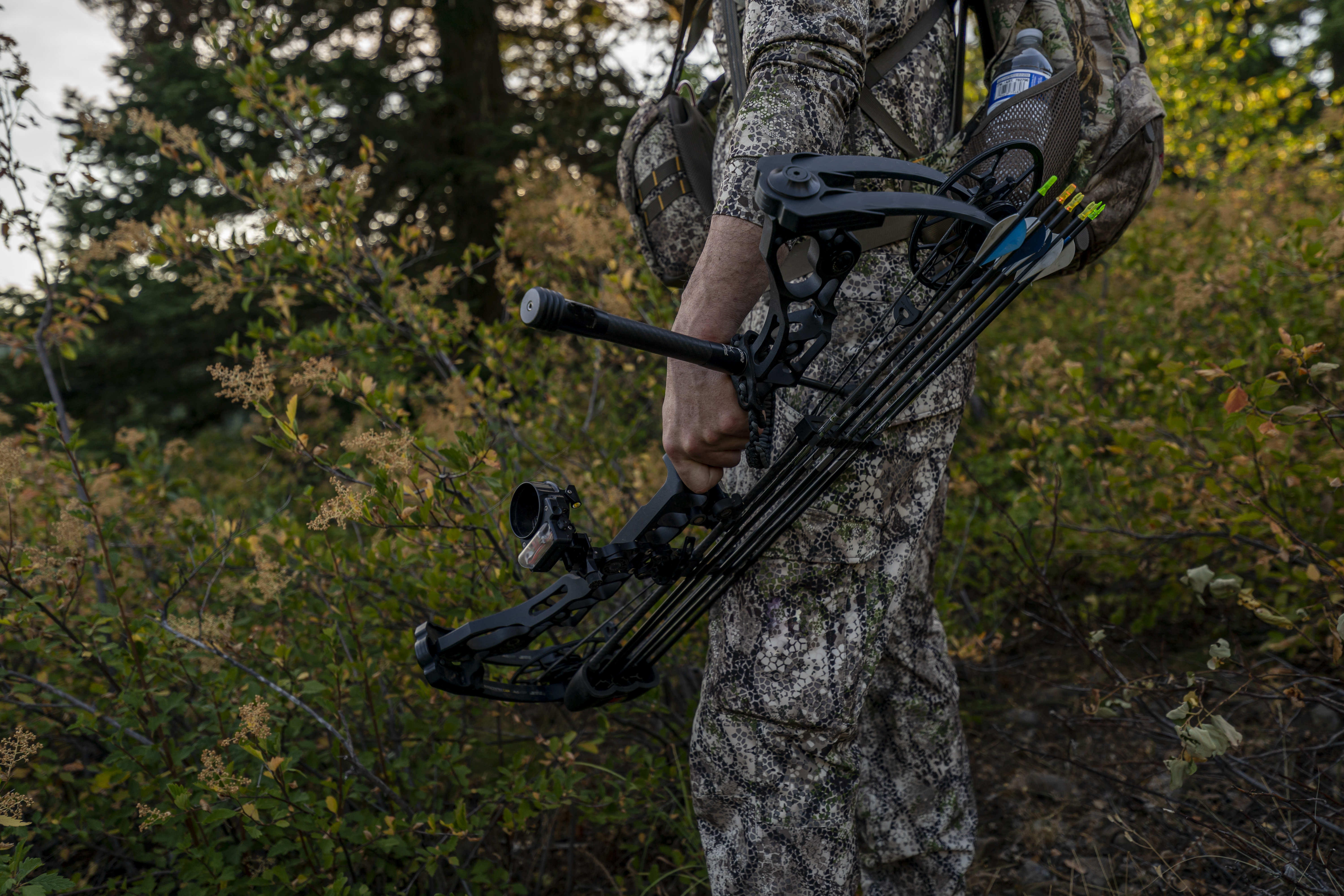 Close up shot of a hunter dressed in camouflage clothing holding a crossbow at his side while hunting elk in a forest at sunset. 